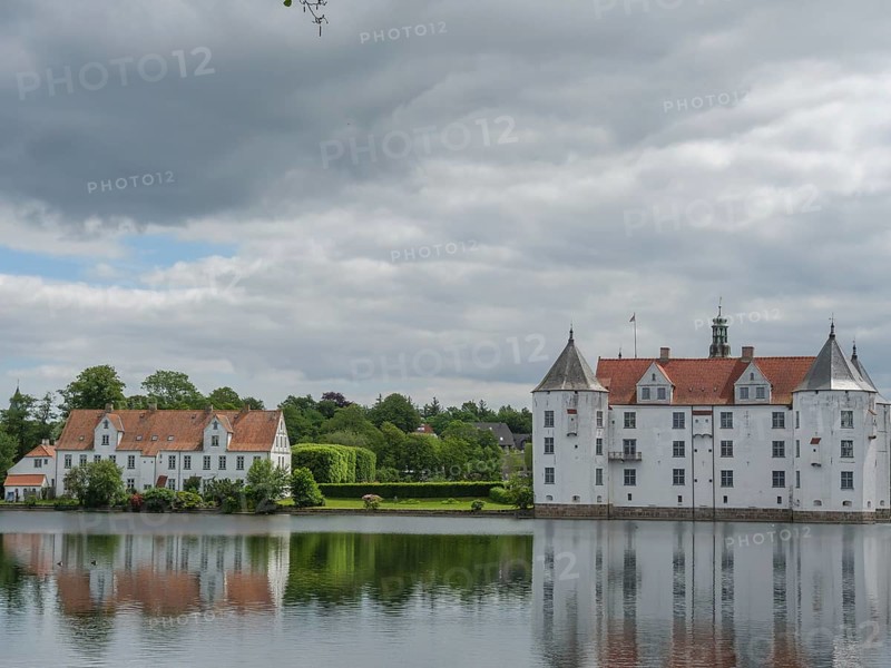 A historic castle with white facades and red roofs is reflected in a calm body of water under a cloudy sky, Glücksburg, Schleswig-Holstein, Germany