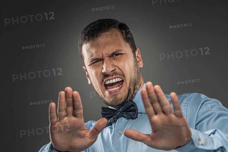 Portrait of angry upset young man in blue shirt and butterfly tie with hands up yelling isolated on gray background. Negative human emotion, hands show Stop, facial expression. Closeup