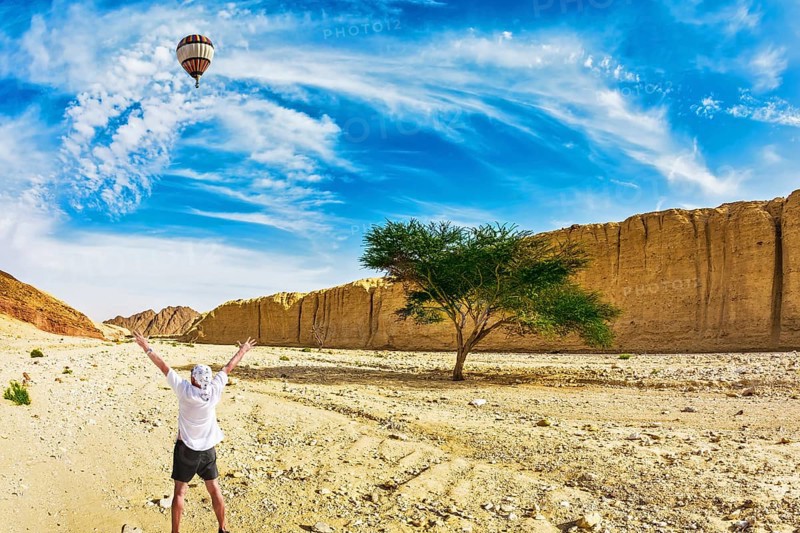 The huge multi-colored balloon flies over desert near the seaside resort of Eilat. The tourist in white t-shirt is delighted with desert