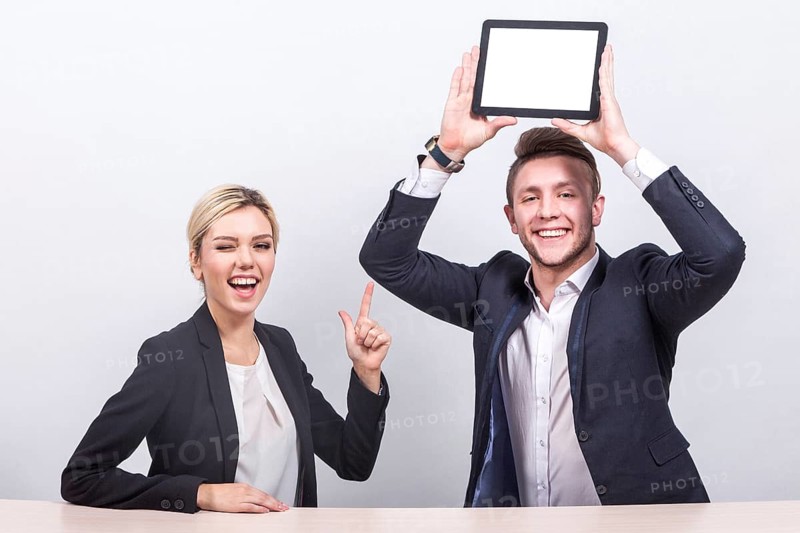 Attractive young caucasian businesswoman with ponytail in black suit and businessman in black suit sit at the office desk and hold empty tablets, cheerful Isolated on grey
