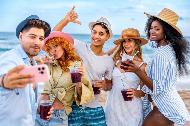 Group of cheerful young multi ethnic friends in summer clothes taking selfie with smartphone and drinking cocktails on the beach