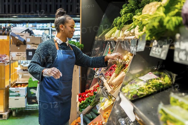 Grocery store employee wearing gloves arranging organic vegetables on display in supermarket