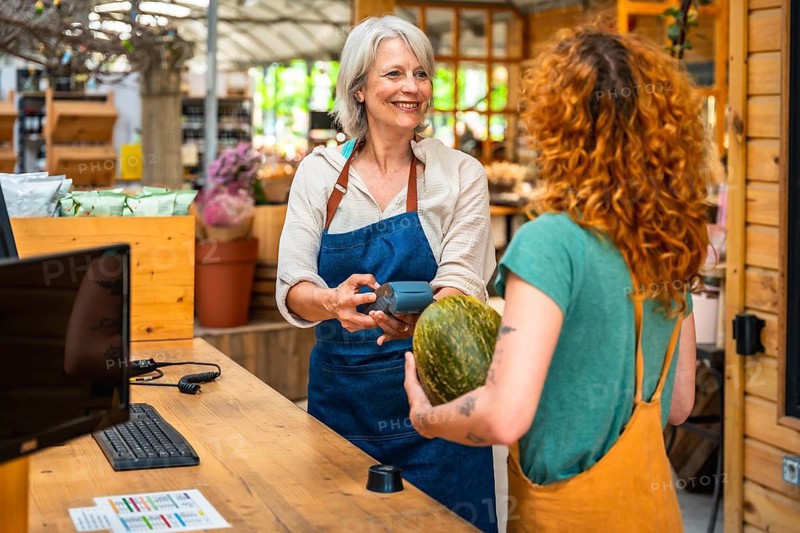 Smiling greengrocer holding pos terminal while customer paying with contactless payment for groceries in a small local shop