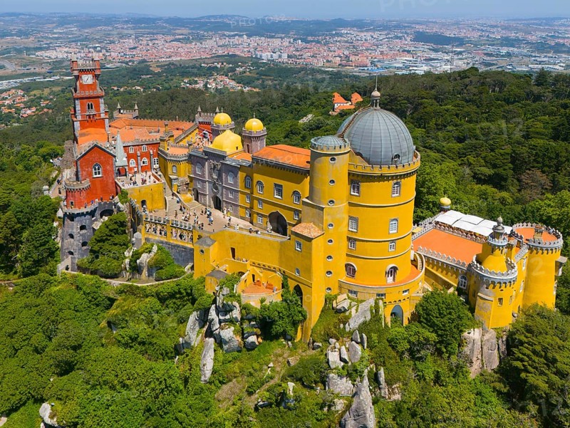 Impressive castle with colourful towers and domed roof on a mountain ridge amidst green trees and wide landscape, aerial view, Palácio Nacional da Pena, National Palace Pena, Sintra, Lisbon, World Heritage Site, UNESCO, Portugal