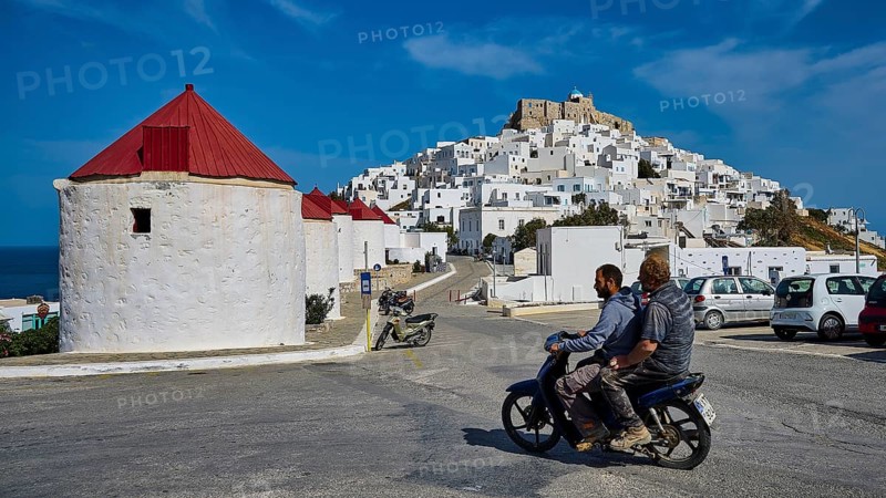 Two people on motorbike in front of white village and windmills on hill, Querini Castle, Chora, Astypalea, Dodecanese, Greek Islands, Greece