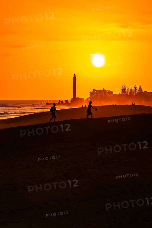 Tourists walking on the maspalomas dunes at sunset, with the maspalomas lighthouse in the background, gran canaria, canary islands, spain