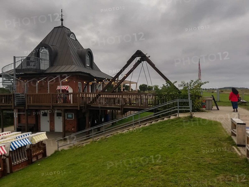 Wooden building with outdoor terrace under a cloudy sky, a person walks past Strandbude Ostseeinsel Fehmarn Fehmarnsund Germany Putgarden