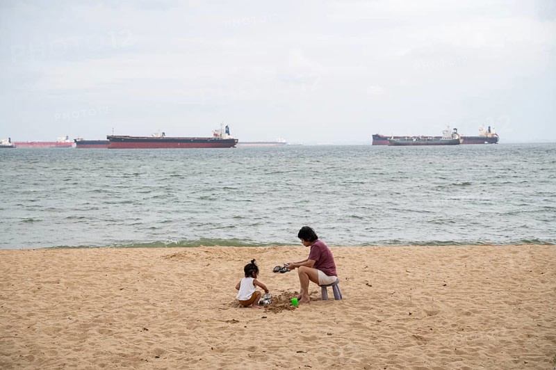 31.07.2023, Singapore, Republic of Singapore, Asia, A woman and a small child sit in the sand on the beach of East Coast Park while container ships can be seen on the water in the background
