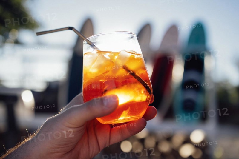 Orange slice and aperol spritz cocktail on a stone against the backdrop of a sandy beach. Man holding slass with iced citrus sparkling mineral water and straw. Copy space