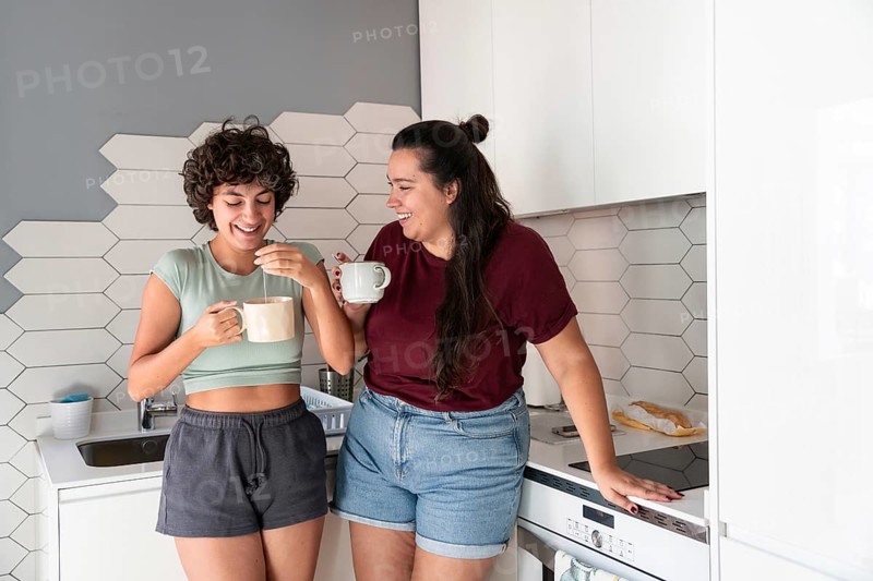 A lesbian couple stands in a cozy kitchen, sipping morning coffee. The modern decor and soft lighting create a warm atmosphere, perfect for a pleasant start to the day