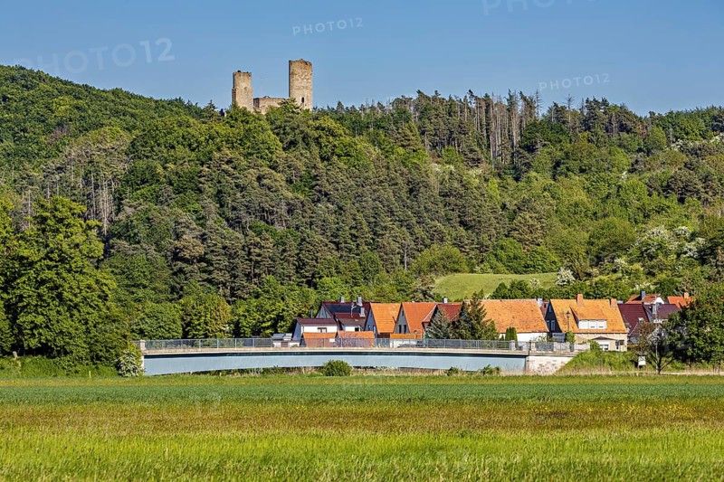 View of a ruined castle above a village with a bridge, surrounded by green forests and meadows under a clear sky, The Brandenburg ruins in the Werra valley near Lauchröden in Thuringia