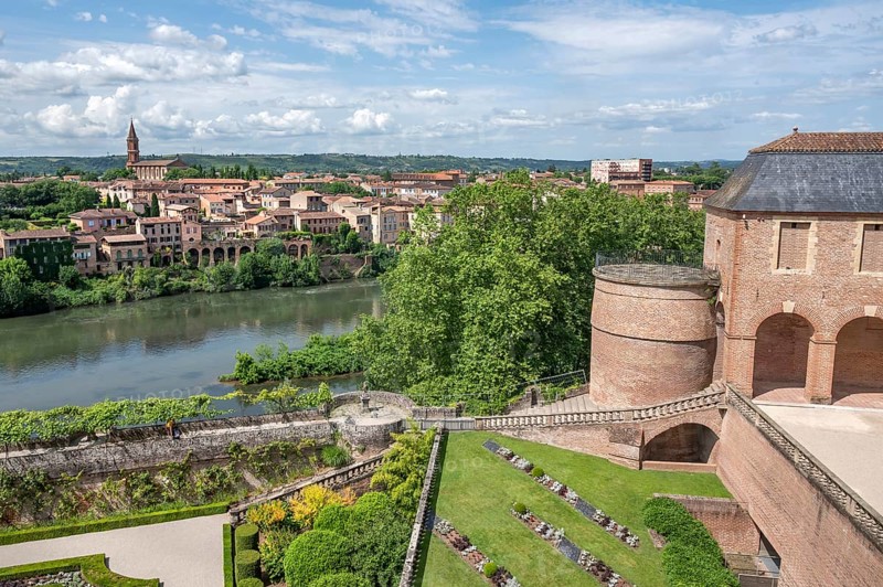 Panoramic view from the Palais de la Berbie across the Tarn to the opposite bank with the striking neo-Gothic Saint-Madeleine church, whose slender bell tower towers over the La Madeleine district, embedded in an ensemble of historic brick houses and lush greenery along the riverbank, characteristic combination of medieval fortress architecture, water landscape and southern French cityscape, Albi, Tarn, Occitanie, France