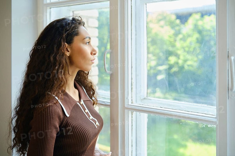 A young woman with long, curly hair is looking out a large window. Bright sunlight fills the room, and the lush greenery outside creates a serene atmosphere. She appears contemplative and calm