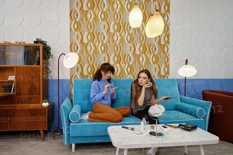 Two young women are engaged in a casual conversation about makeup while sitting on a stylish blue couch in a living room