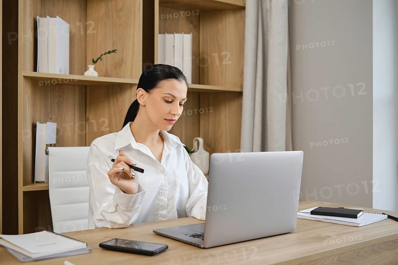 A young woman sits at a clean wooden desk focused on her laptop. She holds a pen in one hand, surrounded by notebooks and a smartphone. The minimalist office features light-colored walls and shelves