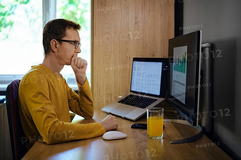 A middle-aged man sits at a wooden desk in a home office, focused on a computer screen displaying data visualisations