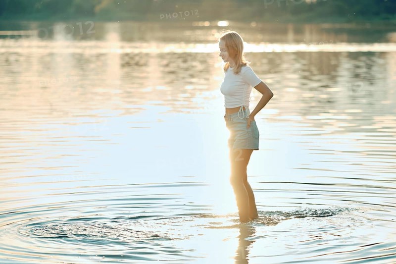 A young woman in a white top and denim shorts stands in shallow water during sunset. The golden sunlight reflects on the water's surface, creating a serene atmosphere as she enjoys the moment