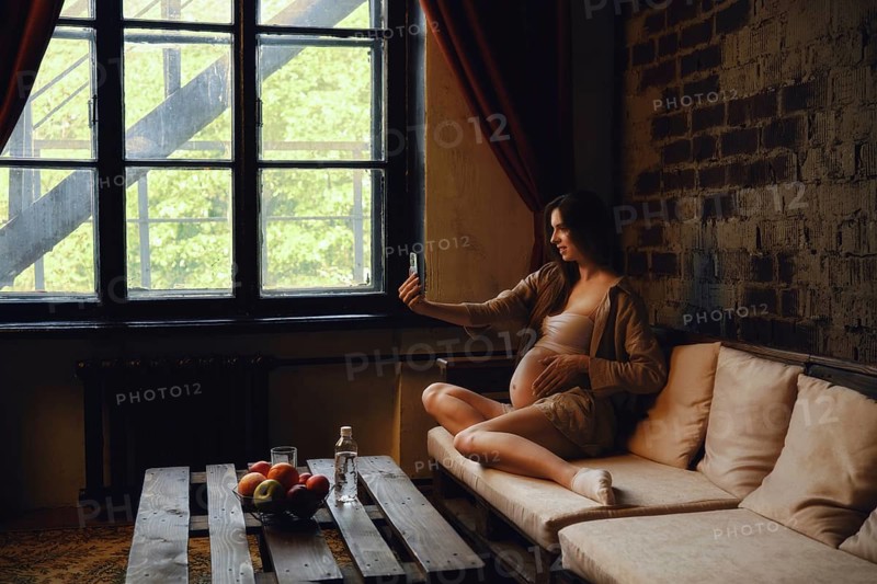 A pregnant woman sits on a comfortable couch at home, casually taking a selfie with her phone. Sunlight streams through the window, illuminating her serene expression