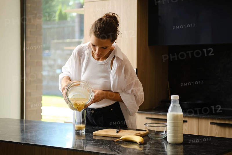 A person blends banana and sea buckthorn in a cozy kitchen. Ingredients are prepared on the counter, with natural light highlighting the freshness of the smoothie-making process