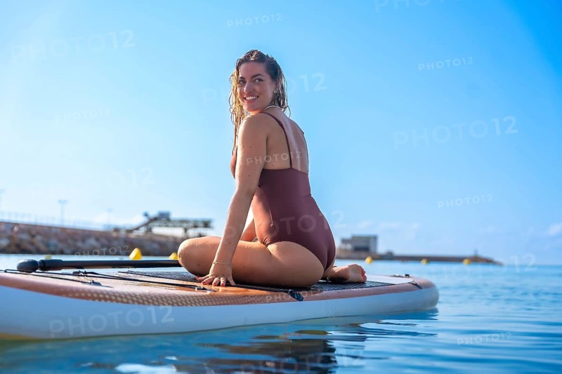 Smiling woman sitting on a paddleboard in calm blue waters, enjoying a sunny summer day at the beach. Clear sky and distant shoreline in the background
