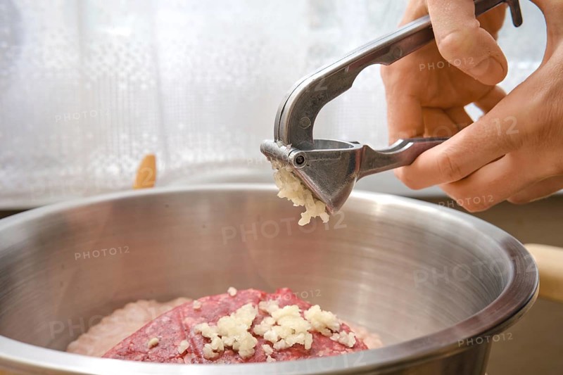 A hand is using a garlic press to add freshly minced garlic to a bowl with ground meat. This cooking preparation takes place in a bright kitchen setting during the day