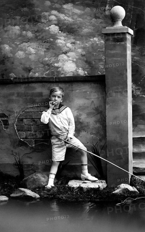 Smoking boy, 1920s, A boy stands fishing at a small pond in front of a painted backdrop, Historical photo