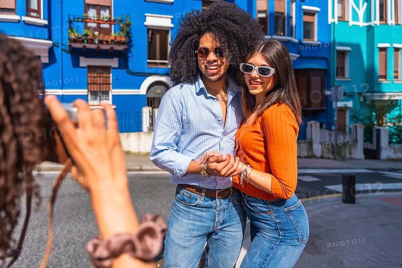 Young photographer taking pictures of a happy tourist couple posing in a colorful street during a sunny day