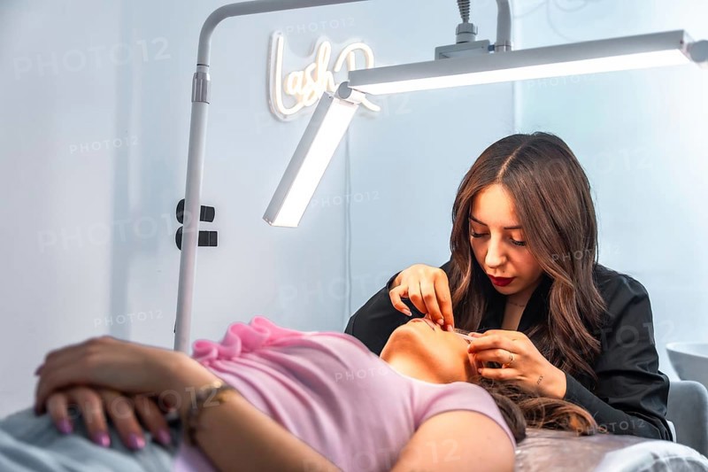 Beautician applying false eyelashes to a client lying on a bed in a beauty salon, illuminated by a professional lamp