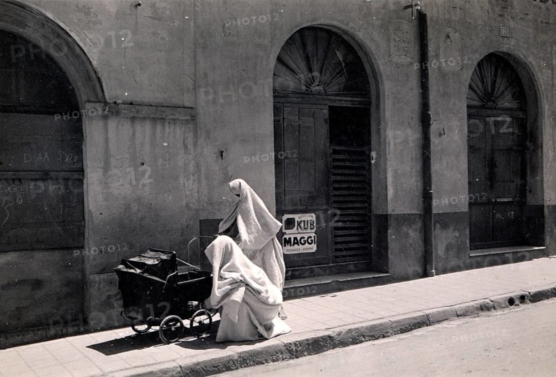 Two Arab women with prams, 1910s, A veiled person sits next to a pram in front of an old building façade, Historical photo