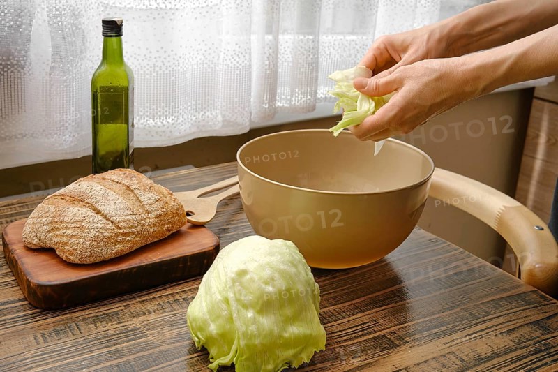 Hands are tearing fresh lettuce over a bowl while a loaf of bread and a bottle of olive oil sit nearby. Sunlight filters through a curtain, creating a warm atmosphere in the kitchen