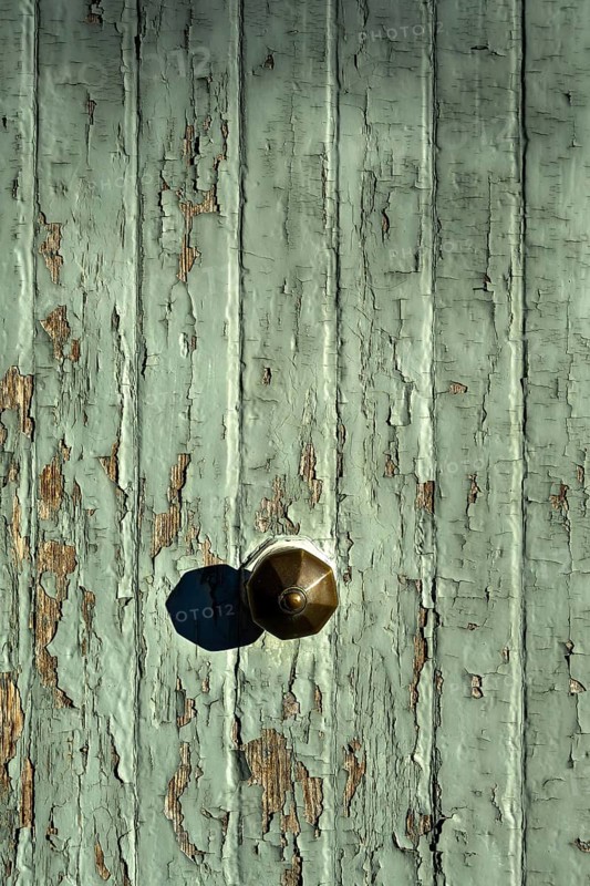 The image captures a close-up of a rustic doorknob on an old, weathered wooden door. The green paint is peeling, revealing the wood beneath, creating a textured and aged appearance