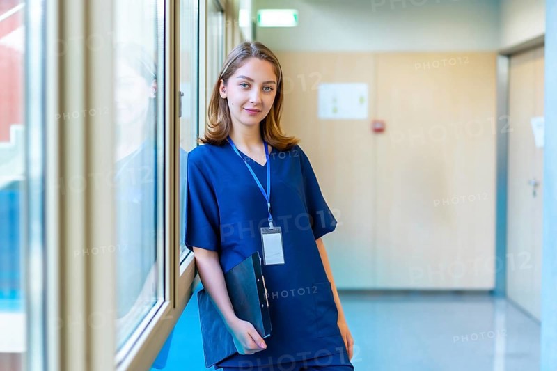 Confident young doctor wearing blue scrubs holding a clipboard while standing in a modern hospital corridor