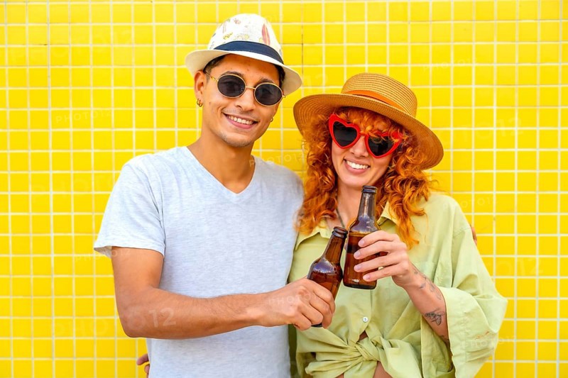 Happy couple toasting with beer bottles while enjoying a summer day against a vibrant yellow tiled wall