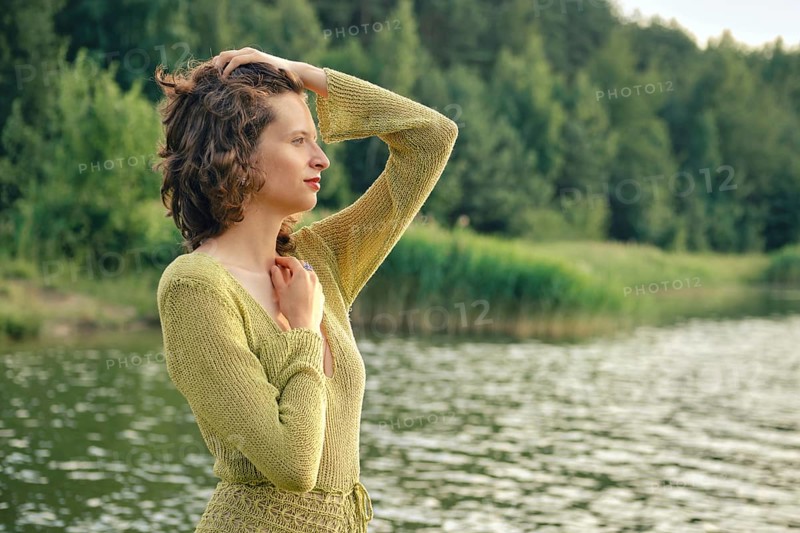 On a hot summer day, a woman stands by the tranquil lake, her hand resting on her head while she gazes thoughtfully at the shimmering water. Lush greenery surrounds the scene, enhancing the beauty