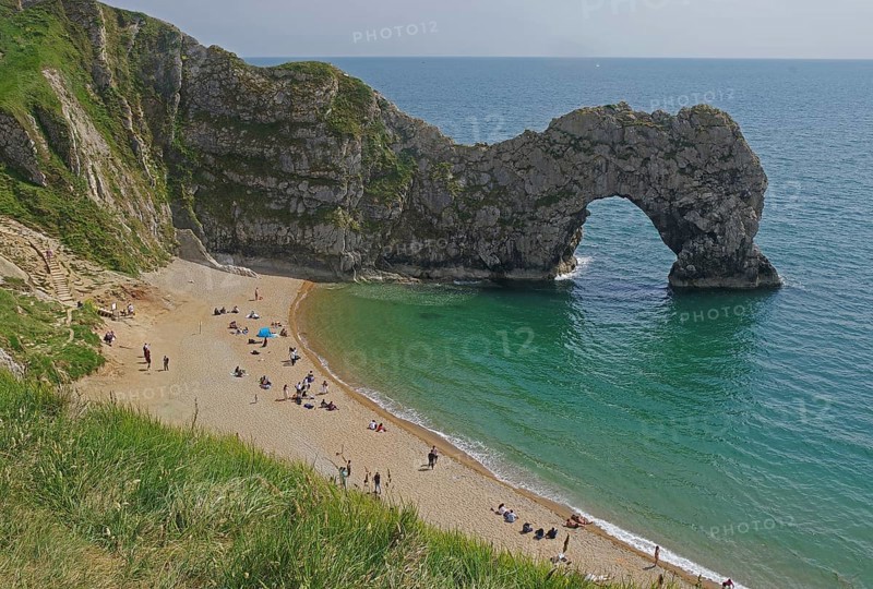 People relaxing on the beach in front of a striking arched cliff and blue sea, Durdle Door, Weymouth, Dorset, England, United Kingdom