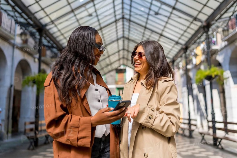 Two cheerful businesswomen are enjoying a moment together, using a smartphone and holding a reusable coffee cup in a covered gallery
