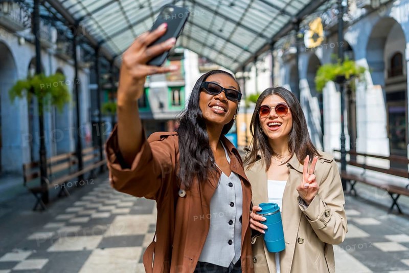 Two female friends taking a selfie in a covered walkway in a city center, one is holding a reusable cup