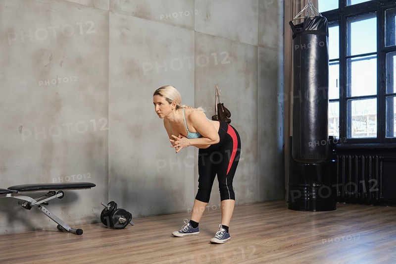 A woman is engaged in a strength training workout at a gym. She is bending forward with her hands clasped, focusing on her form. The room is well-lit, featuring gym equipment