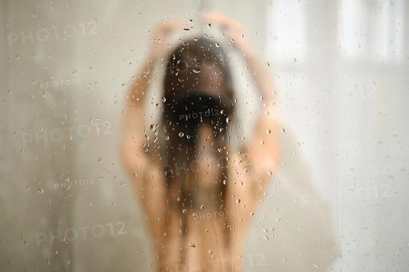 A woman stands in the shower with her hair down, enjoying a peaceful moment. Water cascades down her body, creating a serene atmosphere with droplets gathering on the glass