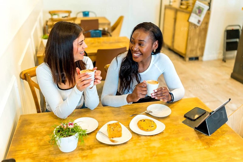 Two female friends enjoying coffee and snacks while chatting and laughing together at a cozy cafe table, sharing a delightful moment