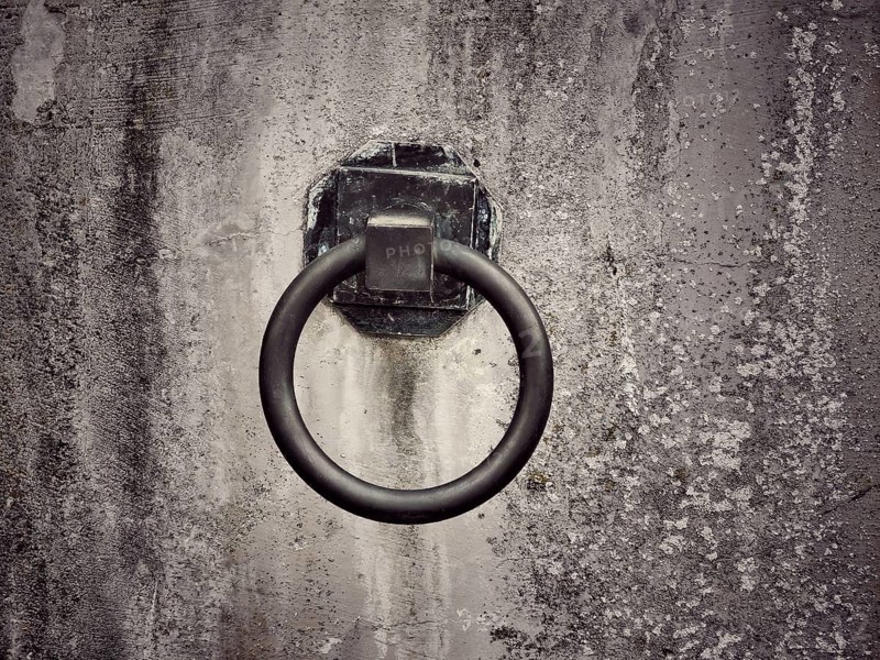 A weathered wall features a rusty metal ring affixed to it, suggesting a historical purpose. The texture and color highlight the passage of time and use in an urban setting