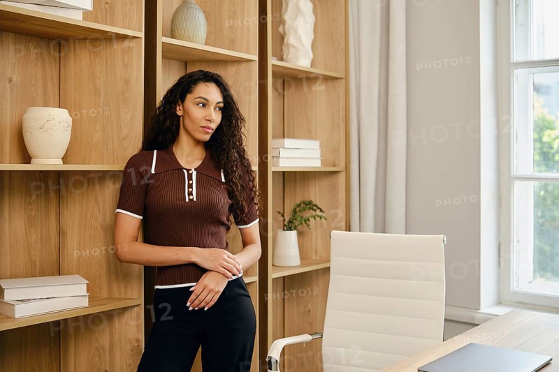 A young woman with long, wavy hair stands in a stylish office. She gazes thoughtfully out of the window, surrounded by wooden shelves, light decor, and natural light adding warmth to the room