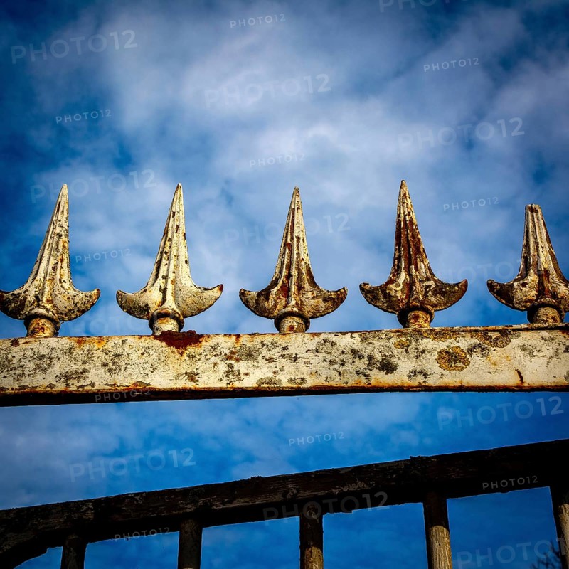 Close-up view of an ornamental iron fence featuring pointed spikes, showcasing rust and weathering against a dramatic cloudy sky in a garden setting. France