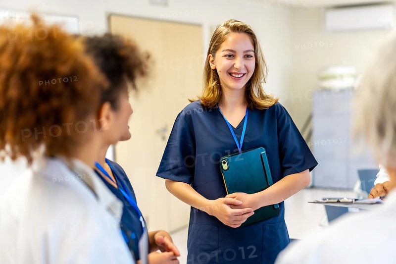 Happy nurse holding a tablet and smiling while engaging in a briefing with the medical team in a bustling hospital corridor
