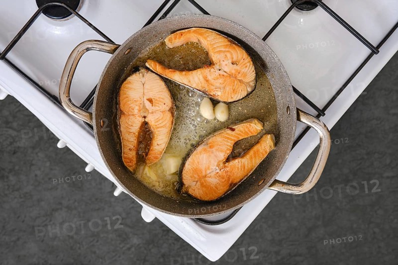 Salmon fillets are being cooked in a pan with garlic on a stovetop. The kitchen is well-lit and the fillets show a golden color, indicating they are almost ready to be served