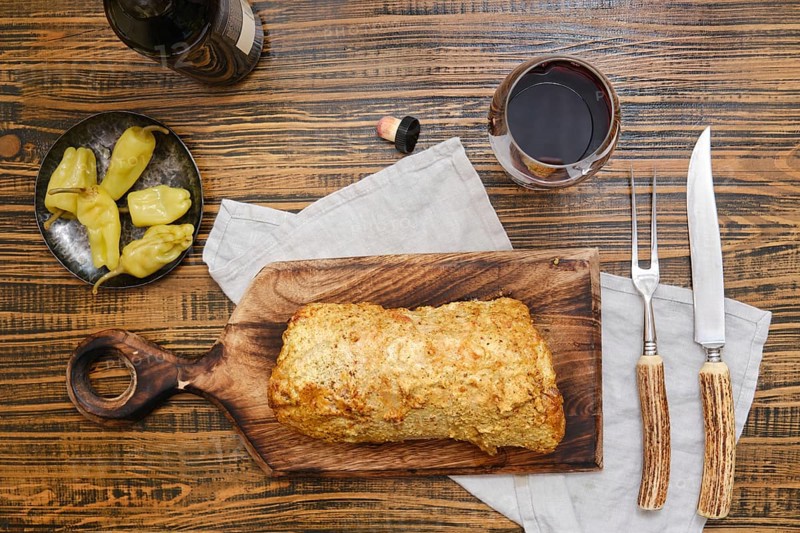 Delicious homemade roasted pork meat and pickled peppers on a rustic wooden table surrounded with red wine in glass, while utensils and a cloth, creating a warm atmosphere for dining