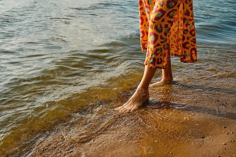 Unrecognizable woman walks barefoot along a sandy beach as waves gently lap at her feet. She wears a vibrant dress with a bold pattern, and the sun sets, casting warm hues on the water