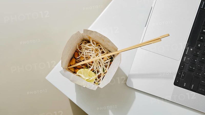 A cardboard bowl of noodles with vegetables and lime sits next to a laptop on a white table. The setting suggests multitasking, combining dining and work in a contemporary environment