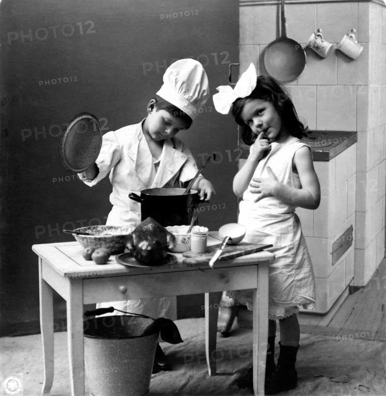 Two children cooking, 1920s, Two children playing in a kitchen, one in a cook's costume, in a black and white photograph, Historical photograph