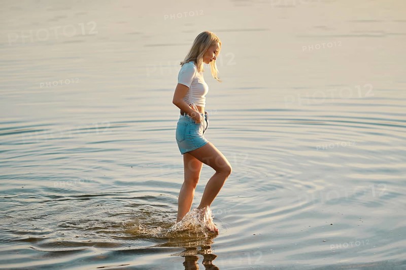 A young woman wearing a white t-shirt and denim shorts walks slowly through the shallow waters of a lake. The sun sets in the background, creating a peaceful atmosphere with gentle ripples